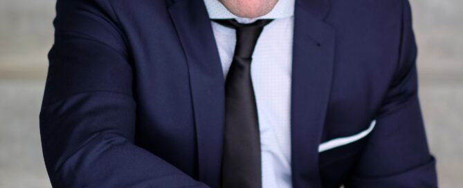 Man in a navy suit and black tie sits on steps, smiling with Corona Notes resting on his knee.