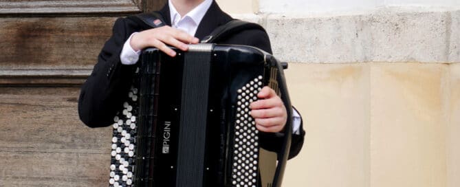 A smiling person in a suit plays a black accordion outdoors, sharing joyful Corona Notes by a wooden door.
