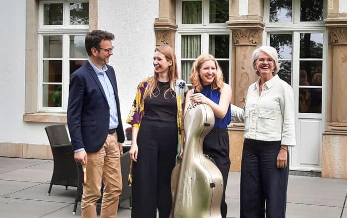 Four adults stand smiling outdoors by a building at meetMUSIC Open Air; one holds a cello case.