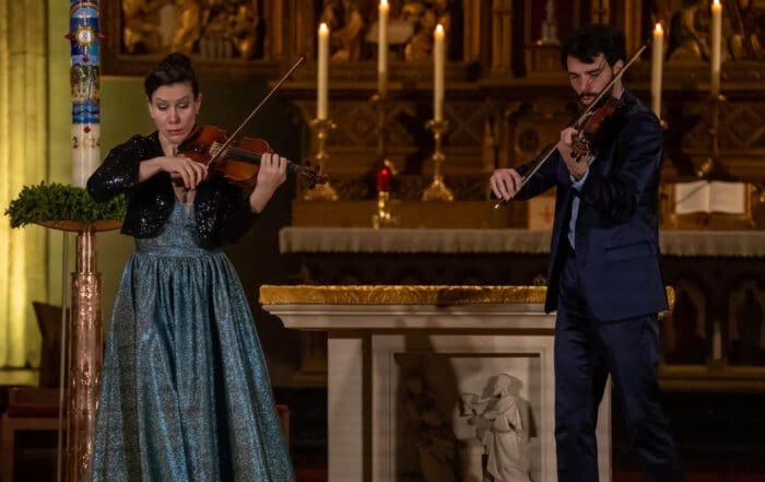 The Twiolins perform in elegant attire inside a church during an Easter Monday concert, with candles glowing behind.