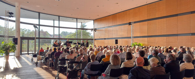 Das Publikum genießt ein Septett beim Summerwinds-Festival in einem sonnendurchfluteten Saal mit großen Fenstern.