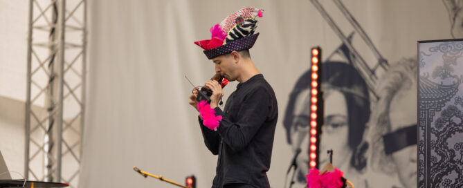 Nico, der einen bunten Hut und eine rosa Boa trägt, hält auf der Bühne des meetMUSIC Open Air eine Rede.
