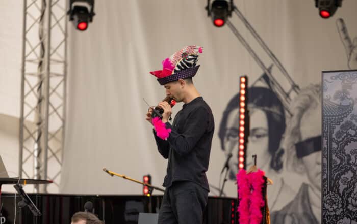 Nico, der einen bunten Hut und eine rosa Boa trägt, hält auf der Bühne des meetMUSIC Open Air eine Rede.