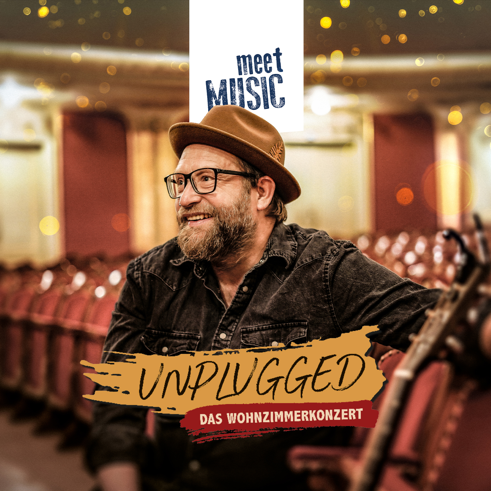 Ein Mann mit Hut und Brille lächelt in einem leeren Theater, hinter ihm sind die Schriftzüge „UNPLUGGED“ und „Gregor Meyle“ zu sehen.